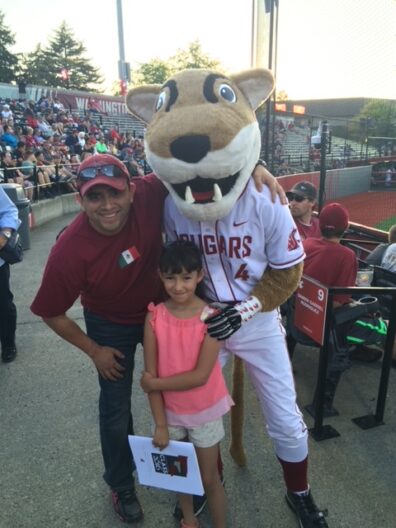 Victor Pimentel posing with a young child beside the WSU Cougar mascot at a crowded baseball stadium, with fans filling the stands behind them.