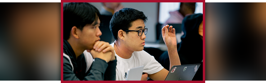 Students seated in a classroom participate in discussion, with one raising a hand beside an open laptop decorated with stickers.