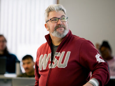 Chris Cooney in a red WSU jacket speaks or gestures while standing in a classroom setting.