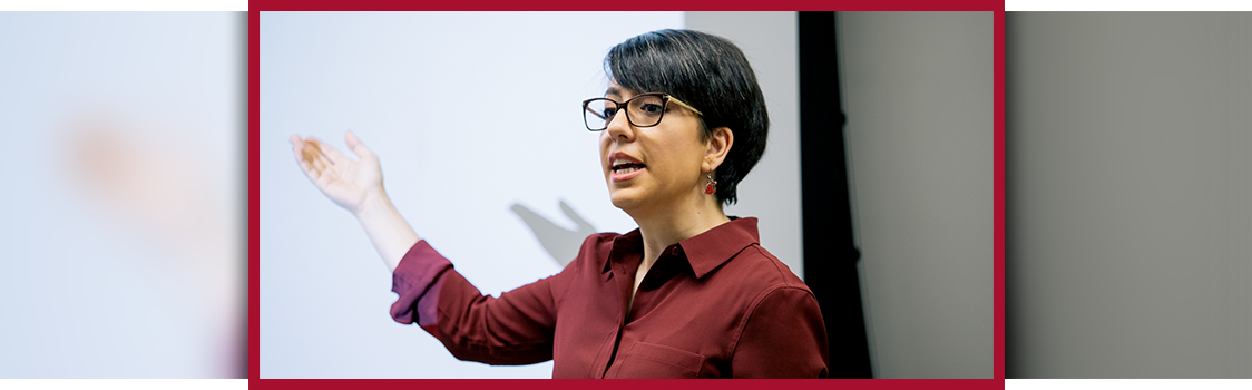 Shirin Shahsavand gestures toward a projection screen while speaking, wearing glasses and a burgundy button‑down shirt in a classroom setting.