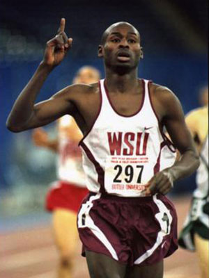 WSU track athlete Bernard Lagat running on an indoor track, wearing a WSU uniform with number 297 and raising one finger.