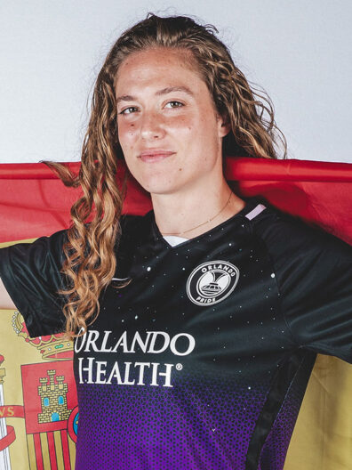 Professional soccer player Celia Jimenez Delgado wearing an Orlando Pride jersey holds a Spanish flag draped behind her.
