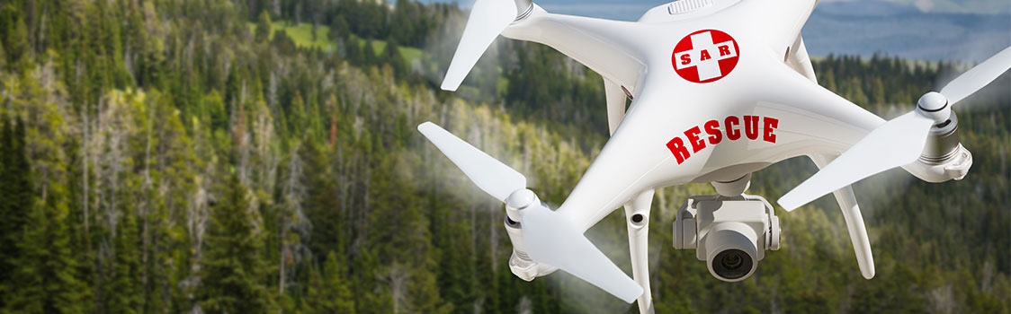 A white quadcopter drone marked with a red “SAR RESCUE” emblem flies above a dense forested landscape with distant mountains under a cloudy sky.