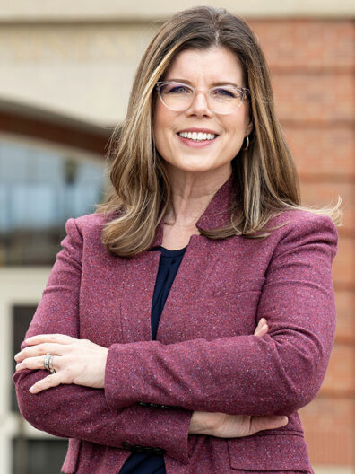 Cheryl Oliver stands outdoors with arms crossed, wearing a maroon blazer over a dark top in front of a brick and glass building.