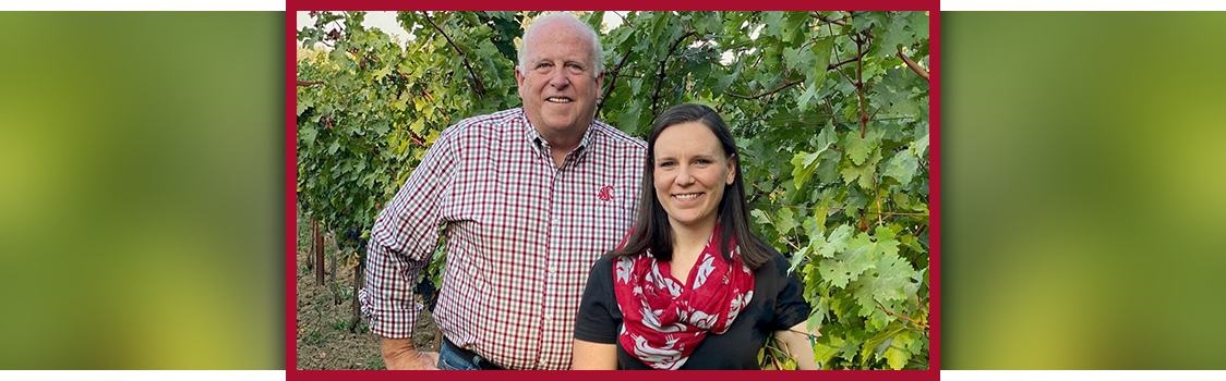 Boyd Family Vineyards owner Stan Boyd and Carter (Marks) Wiggins stand together in front of lush grapevines, posing between rows of green foliage.
