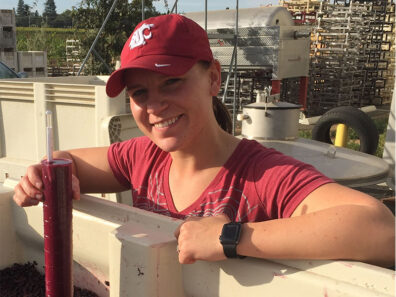 Carter (Marks) Wiggins stands beside a fermentation bin at a vineyard, wearing a WSU cap and red shirt while measuring freshly crushed grapes.