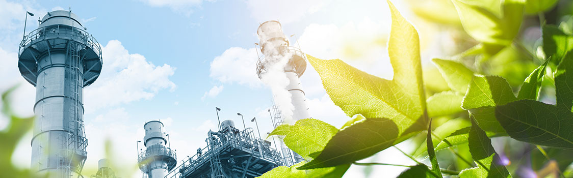 Industrial towers and metal structures rise behind bright green leaves illuminated by sunlight, contrasting manufacturing with natural greenery.