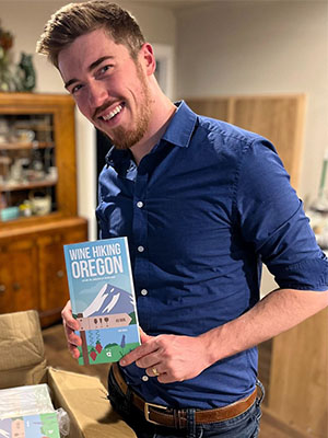 Jack Costa holds a copy of his book “Wine Hiking in Oregon” while standing beside a box of additional copies.