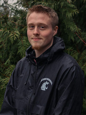 Jagger Norris wearing a rain‑soaked WSU Mechanical Engineering jacket standing outdoors in front of evergreen branches.