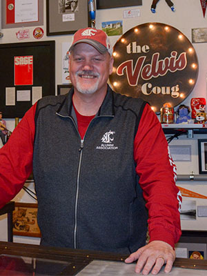 Mark Schuster stands behind a counter in a room decorated with WSU memorabilia, including a sign reading “The Velvis Coug.”