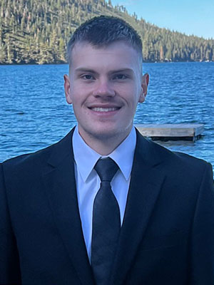 Tyler Shaffer stands by a mountain lake wearing a black suit and tie.