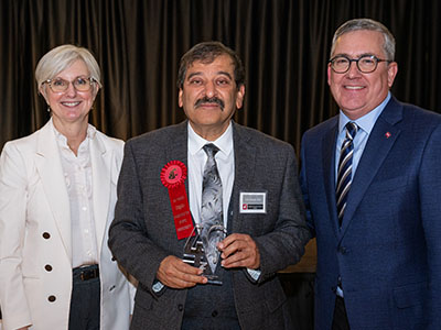 Chancellor Elizabeth Chilton, Umesh, and President Kirk Schulz stand together at an event, with the Umesh in the center holding a service award ribbon and trophy, posed against a dark curtain backdrop.