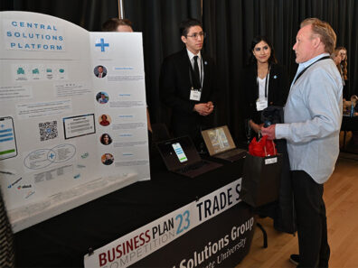 Central Solutions Group team members stand at their trade show booth, presenting a display board and laptop to an attendee during the Business Plan Competition.