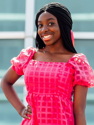 Felicia Adesope stands outdoors wearing a bright pink, textured dress with puff sleeves, one hand on her hip against a soft, modern building backdrop.