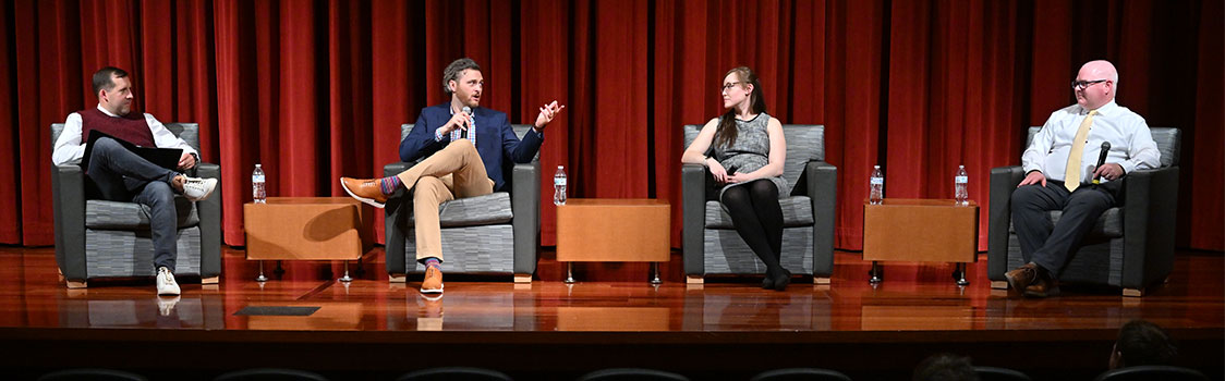 A panel of four speakers, including Professor Robert Crossler (left) with Andrew Pirie, Jasmyn Bearly, and Jason Mayer, sits on stage in armchairs discussing the 2023 Business Technology Symposium.