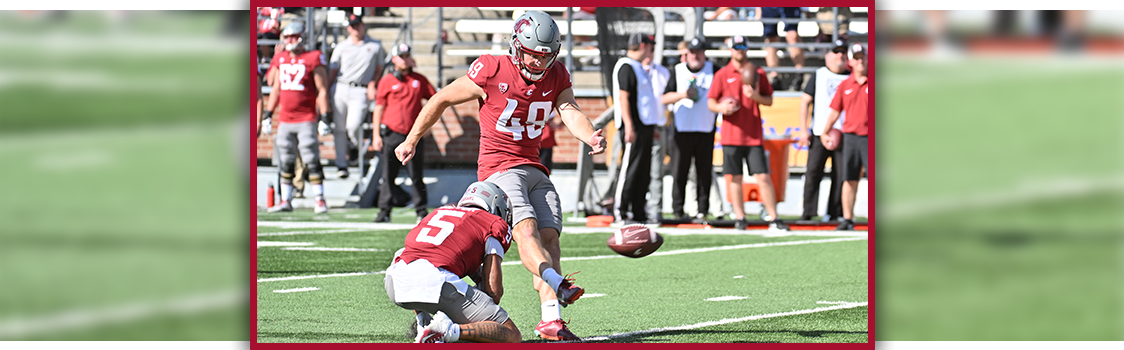 WSU Kicker Dean Janikowski in uniform kicking a football during a game, with a teammate holding the ball and players and coaches standing along the sideline.