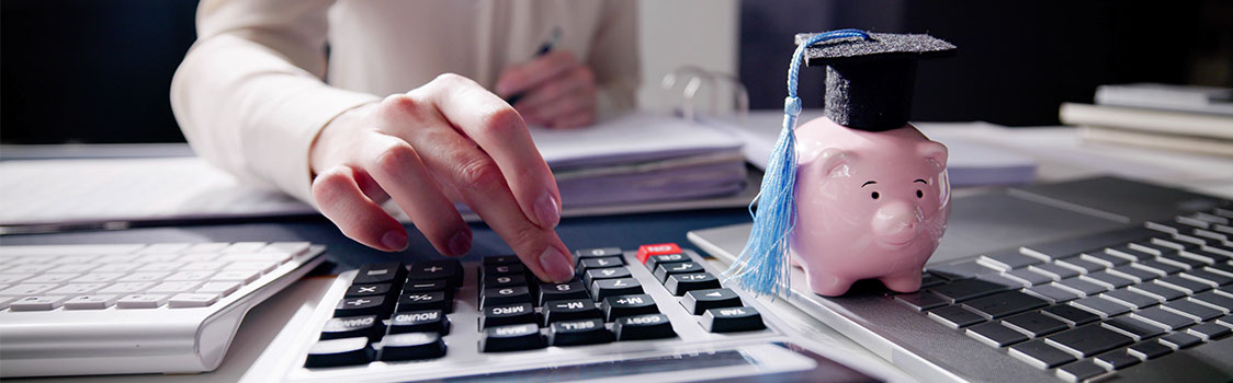 A piggy bank wearing a graduation cap sits atop a stack of documents while a person works with papers and a calculator in the background.