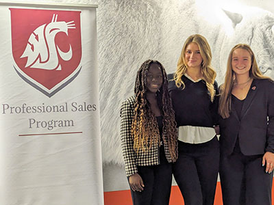 Sylvia Johns, Maya Busby, and Ashley Buhner stand together near a Professional Sales Program banner after the Speed Selling Competition.