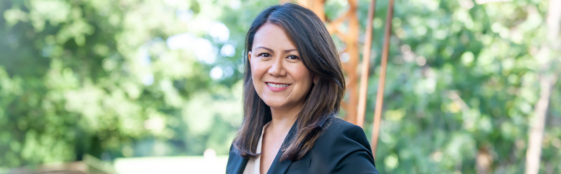 Tina Zingmark stands outdoors in a black blazer, with sunlight filtering through green trees in the background.