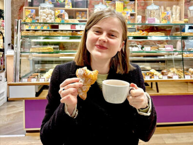 Anika Wottreng sitting in a French bakery, holding a croissant in one hand and a cup of coffee in the other, with pastry displays behind her.