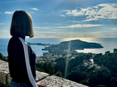Anika Wottreng standing at a stone overlook on the French Riviera, gazing toward the sunlit Cap d’Antibes peninsula and the coastline below.