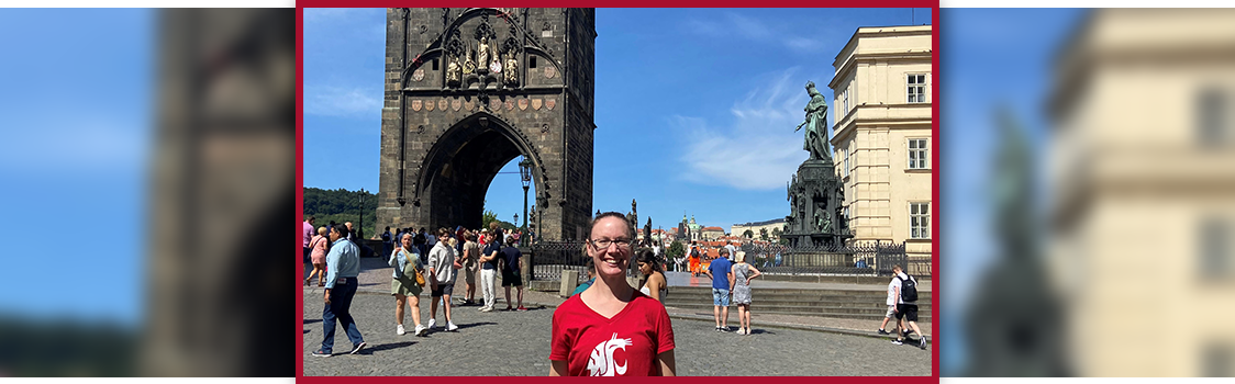 Denise Thrush standing in a red WSU shirt before the entrance tower of the Charles Bridge in Prague, with tourists walking across the cobblestone plaza.