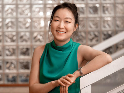 Cathy Jun wearing a sleeveless green top, leaning casually on a white railing in front of a wall made of glass blocks.