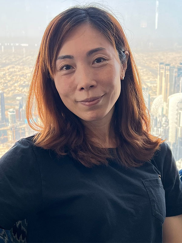 Rachel Tsang in a dark tee with shoulder‑length brown hair before a hazy Hong Kong high‑rise skyline, indoors near a window.