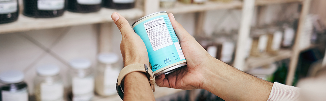 Hands holding a light‑blue canned product, examining its nutrition label in a shop with jars and containers on shelves in the background.
