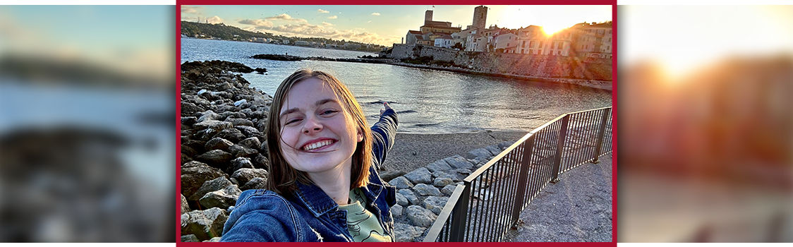 Anika Wottreng extending an arm toward the Antibes coastline at sunset, standing on a rocky path beside calm water and the old town’s sunlit buildings.