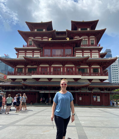 Denise Thrush standing in front of the multi‑tiered Buddha Tooth Relic Temple in Singapore, with visitors walking across the open courtyard below a partly cloudy sky.