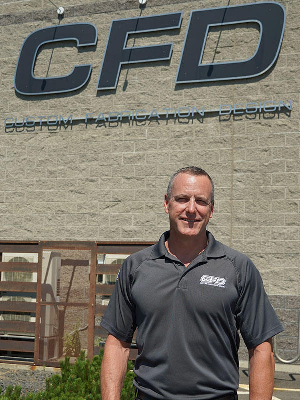 Bob Wiese standing outside the Custom Fabrication Design building, with the CFD logo visible on the gray exterior.