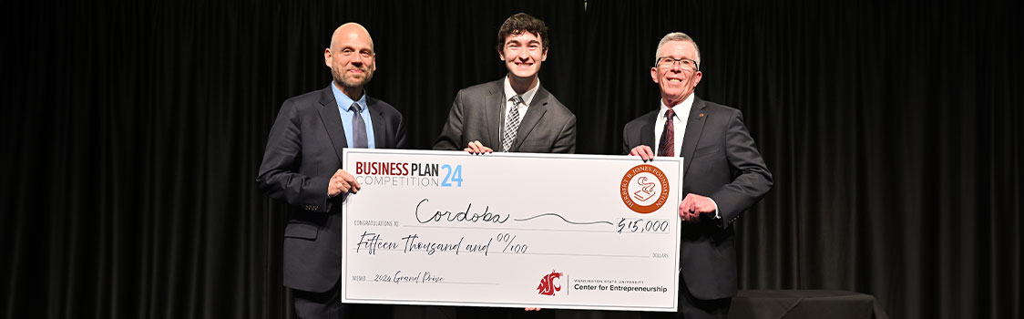 Three individuals holding an oversized Business Plan Competition check for the Cordoba team, celebrating their first‑place $15,000 award onstage.