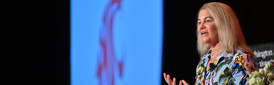 Starbucks CFO Rachel Ruggeri presenting onstage at the 2024 fall Power Breakfast, gesturing while standing before a large screen with the WSU logo.
