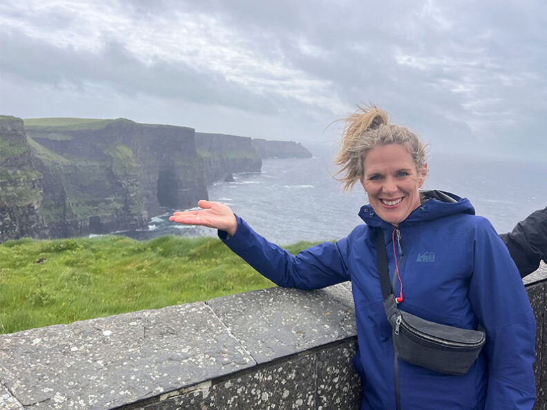 Kay Meyer in a blue rain jacket stands at a stone overlook, gesturing toward the Cliffs of Moher rising above the Atlantic Ocean in County Clare, Ireland.