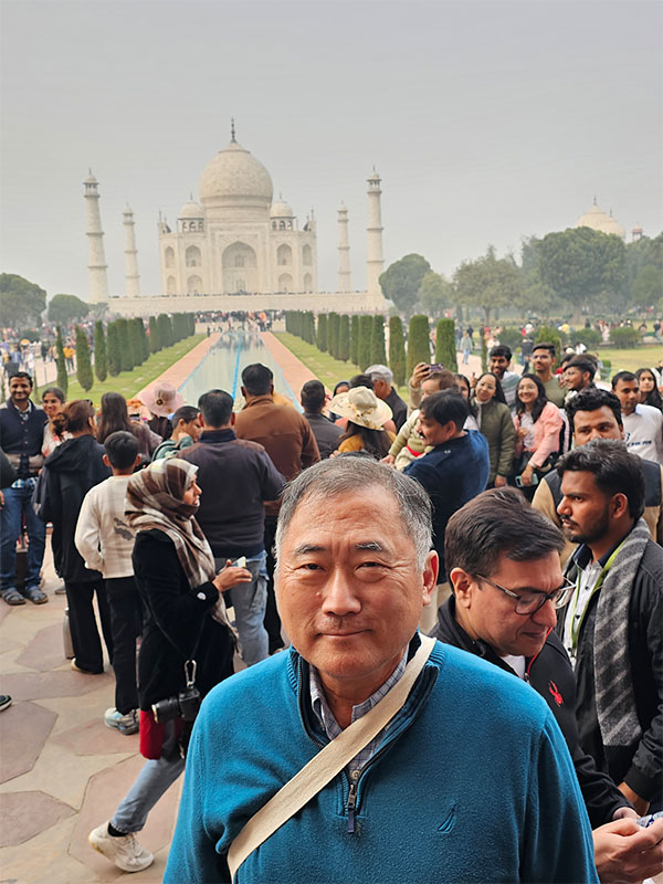 Sung Ahn stands among visitors at the Taj Mahal in Agra, India, with the monument and its long reflecting pool visible in the background.
