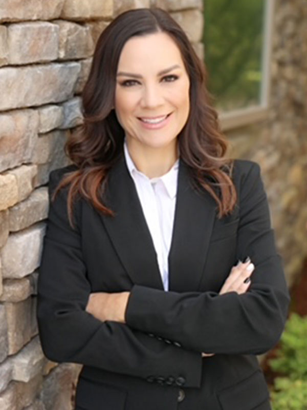 Katie Elliott in a black suit jacket stands with arms crossed against a stone wall, with soft outdoor greenery in the background.