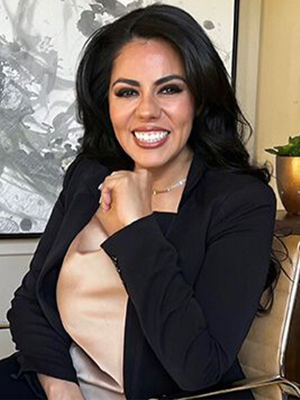 Paulina Gastelum sits in an office chair wearing a black blazer and light blouse, posed with one hand resting near the chin in a modern workspace.