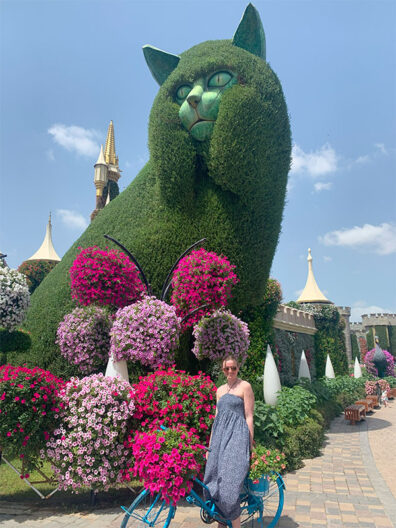 Mary Russell stands beside a flower‑covered bicycle in front of a large green botanical cat sculpture surrounded by vibrant blooms at Dubai’s Miracle Garden.