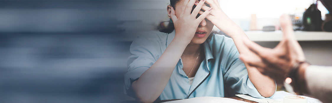 Two people sit across from each other at a desk, with one appearing stressed and the other gesturing during a conversation in a workplace setting.