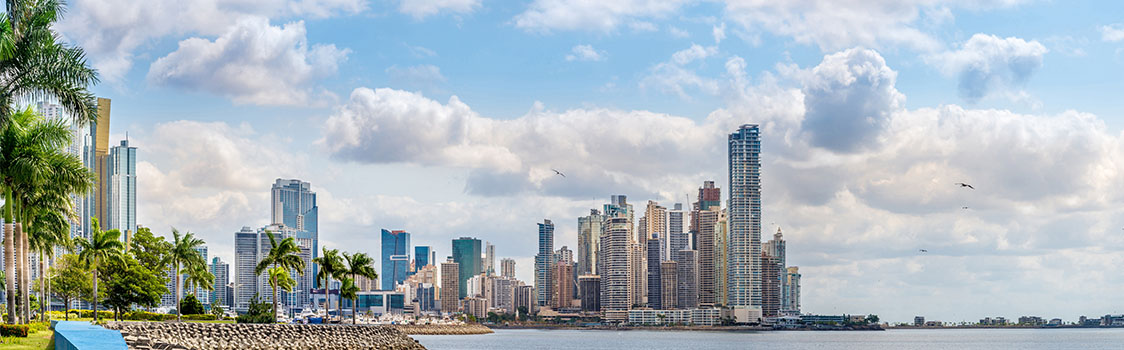 A panoramic view of Panama City’s waterfront skyline with tall modern buildings, palm trees along the shore, and clouds reflected over the bay.