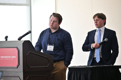 Mason Pickering (left) in a dark blue shirt and khaki pants, and Ben Kapsandy (right) in a suit with a light blue tie, standing in front of a podium labeled 'Washington State University,' listening to feedback from judges.