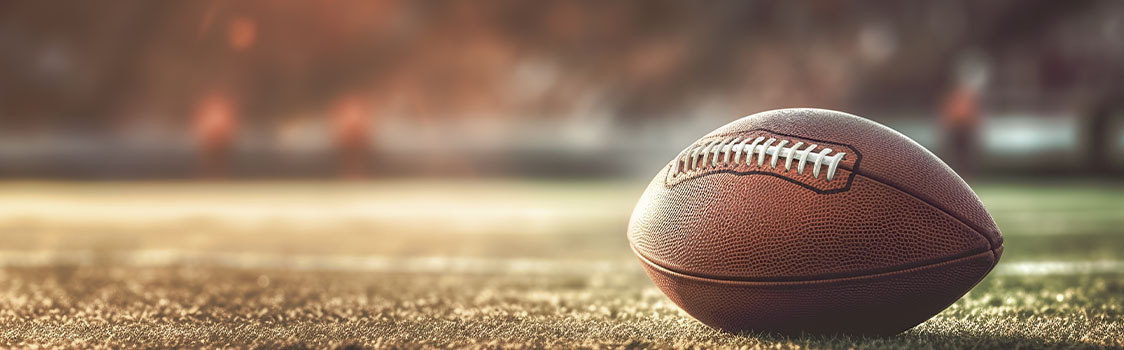 American football resting on a grassy field with a blurred stadium background.