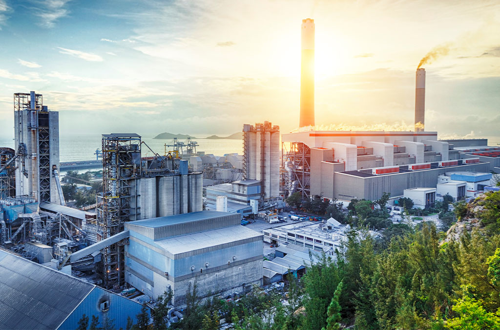 Industrial complex with smokestacks at sunrise or sunset, with trees in the foreground and water in the distance.