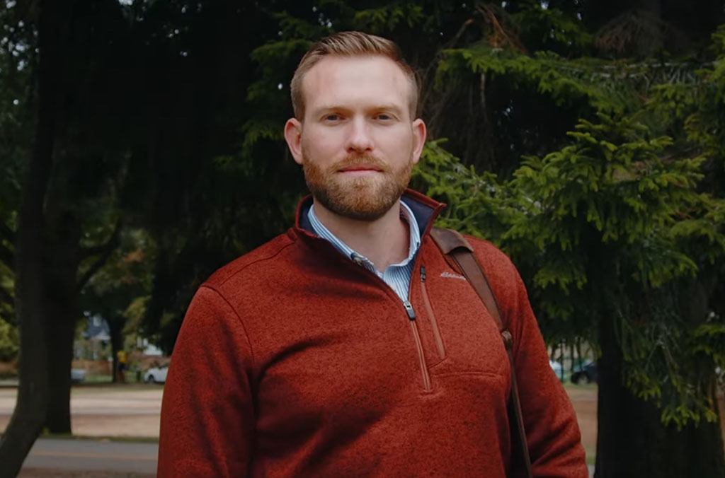 Nick Hill wearing a red quarter-zip sweater and blue collared shirt, standing outdoors with trees in the background.