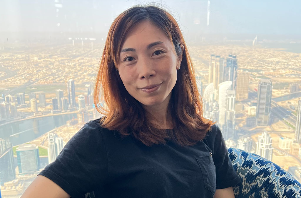 Rachel Tsang in a black shirt seated on a patterned chair with Dubai’s skyline in the background.