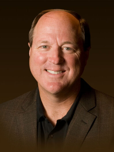 Dan Castles wearing a dark brown blazer over a black collared shirt, set against a solid brown background.