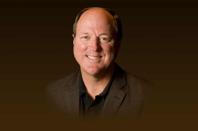 Dan Castles wearing a dark brown blazer over a black collared shirt, set against a solid brown background.