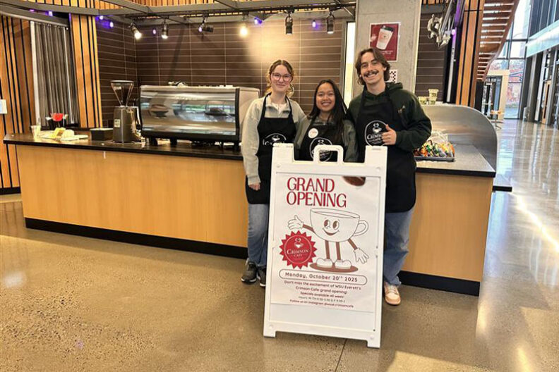 Three students wearing Crimson Café aprons standing behind a grand opening sign inside the new student-run coffee shop at WSU Everett, with a counter and espresso machine in the background.
