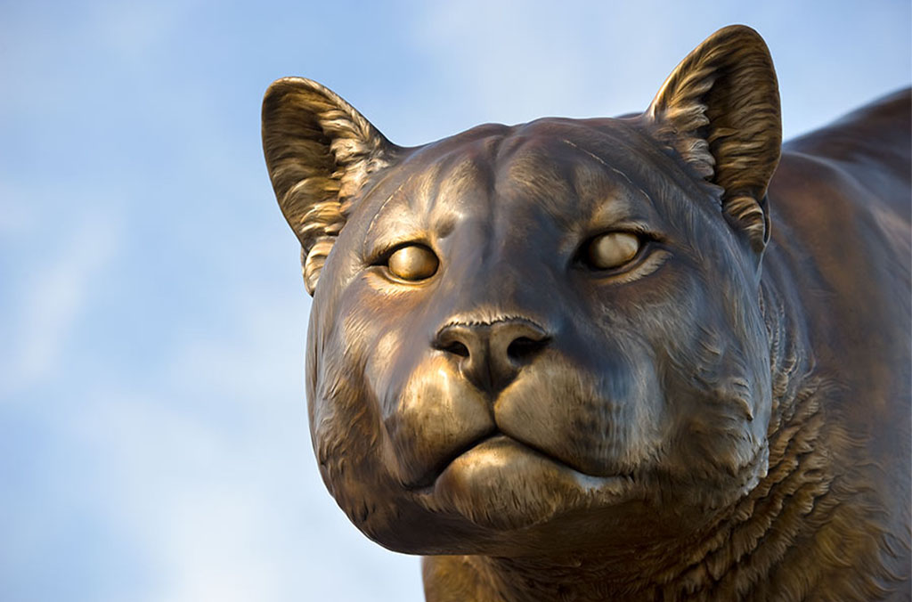 Close-up of a bronze cougar statue with detailed ears against a blue sky.